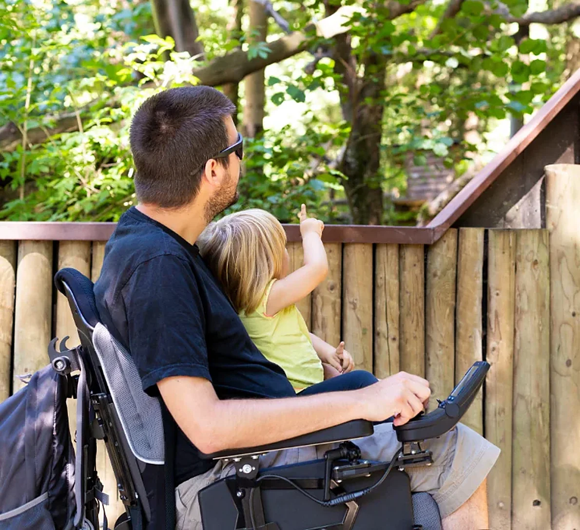 A father seated in a wheelchair and a young child looking out together from a wooden deck.