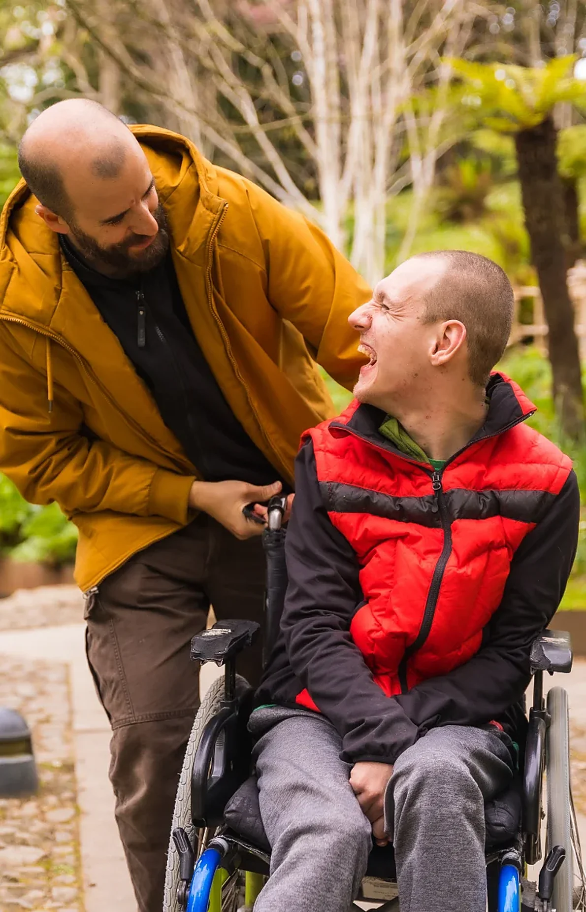 Two friends laughing together outdoors, one of them seated in a wheelchair.